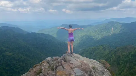 Francesca Murphy Francesca Murphy standing on top of the mountain looking at the beautiful view of green, lush mountains. Back to the camera and he holds out his hand and takes in the view. 