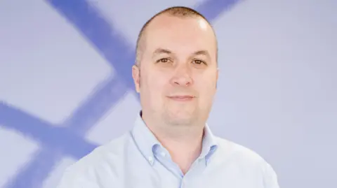 Mind Cymru Simon Jones, a white man with shaved brown/grey hair looks into the camera, smiling slightly. He wears a light blue shirt and stands in front of a white and blue background.