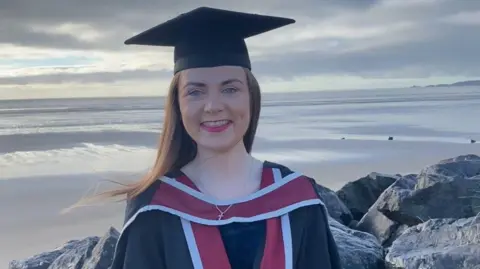 Francesca Murphy Francesca Murphy wearing her graduation gown and cap, standing, looking into the camera with Swansea beach behind her. She smiled.